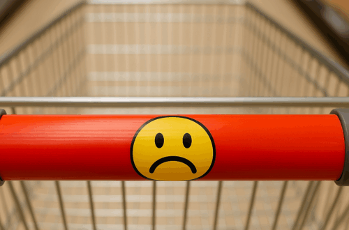 Einkaufswagen mit rotem Griff und traurigem Smiley-Aufkleber im Supermarktgang