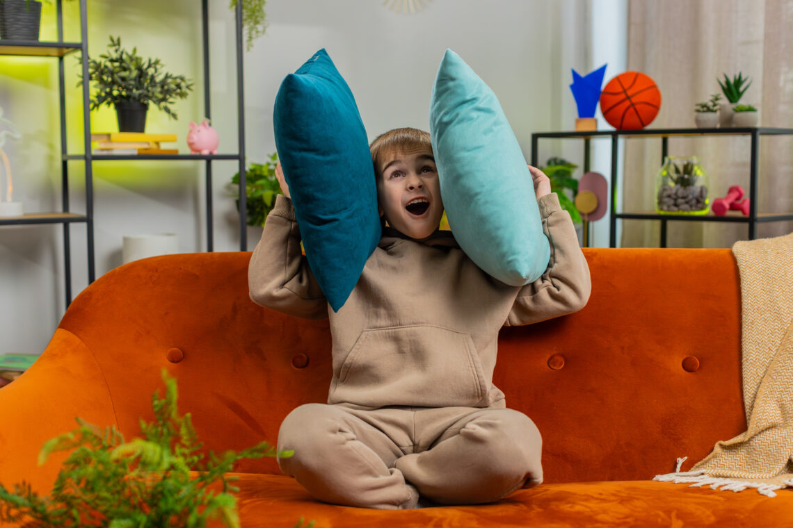 Kind sitzt auf einem orangefarbenen Sofa und hält sich zwei Kissen an die Ohren, als wolle es laute Musik oder Lärm ausblenden.