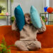 Kind sitzt auf einem orangefarbenen Sofa und hält sich zwei Kissen an die Ohren, als wolle es laute Musik oder Lärm ausblenden.