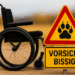 Ein leerer Rollstuhl steht auf einem Feldweg, daneben ein gelbes Warnschild mit der Aufschrift „Vorsicht bissig!“ und einer stilisierten Pfote